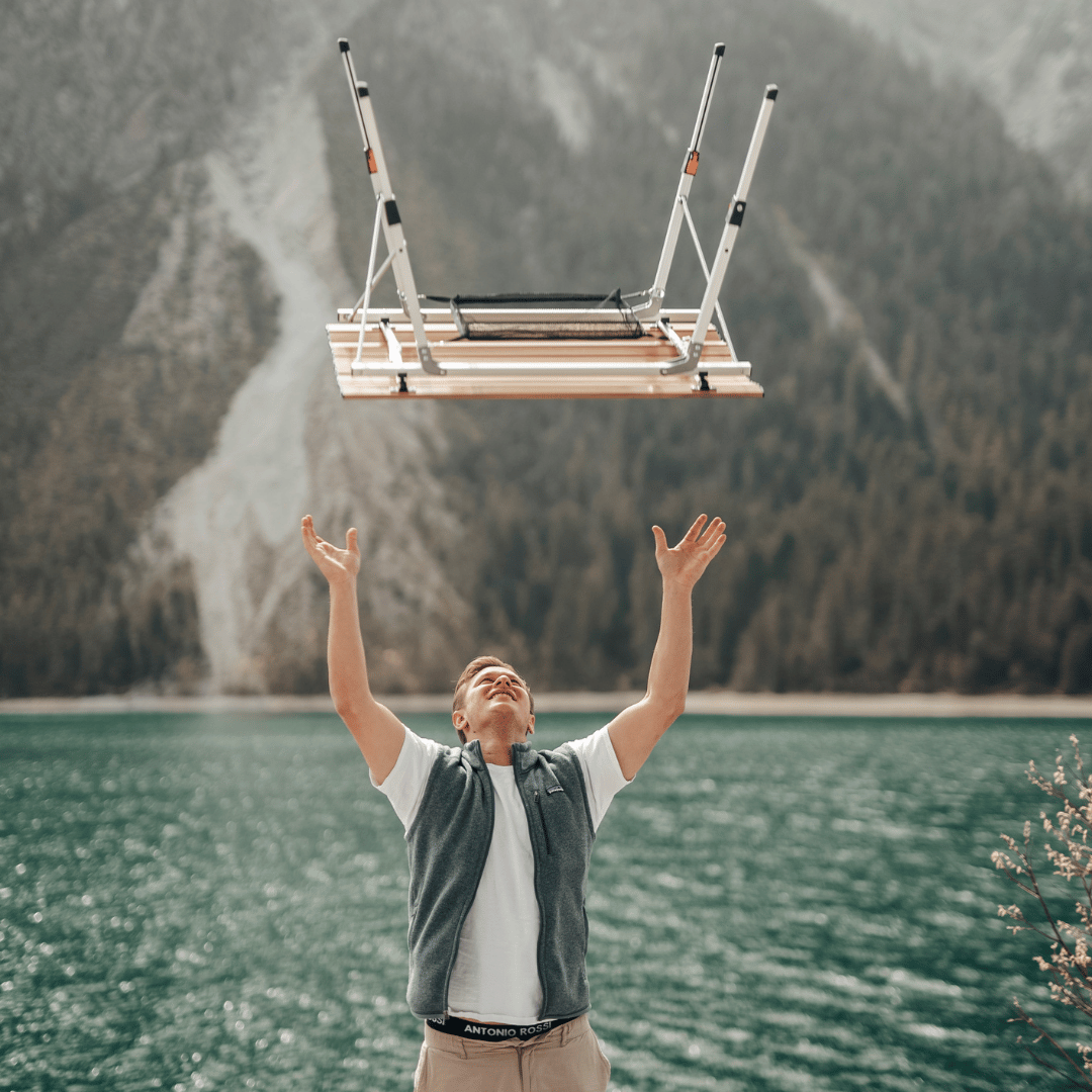 Ein Mann mit einer grünen Weste wirft einen Campingtisch in die Luft. Im Hintergrund befindet sich ein Bergsee