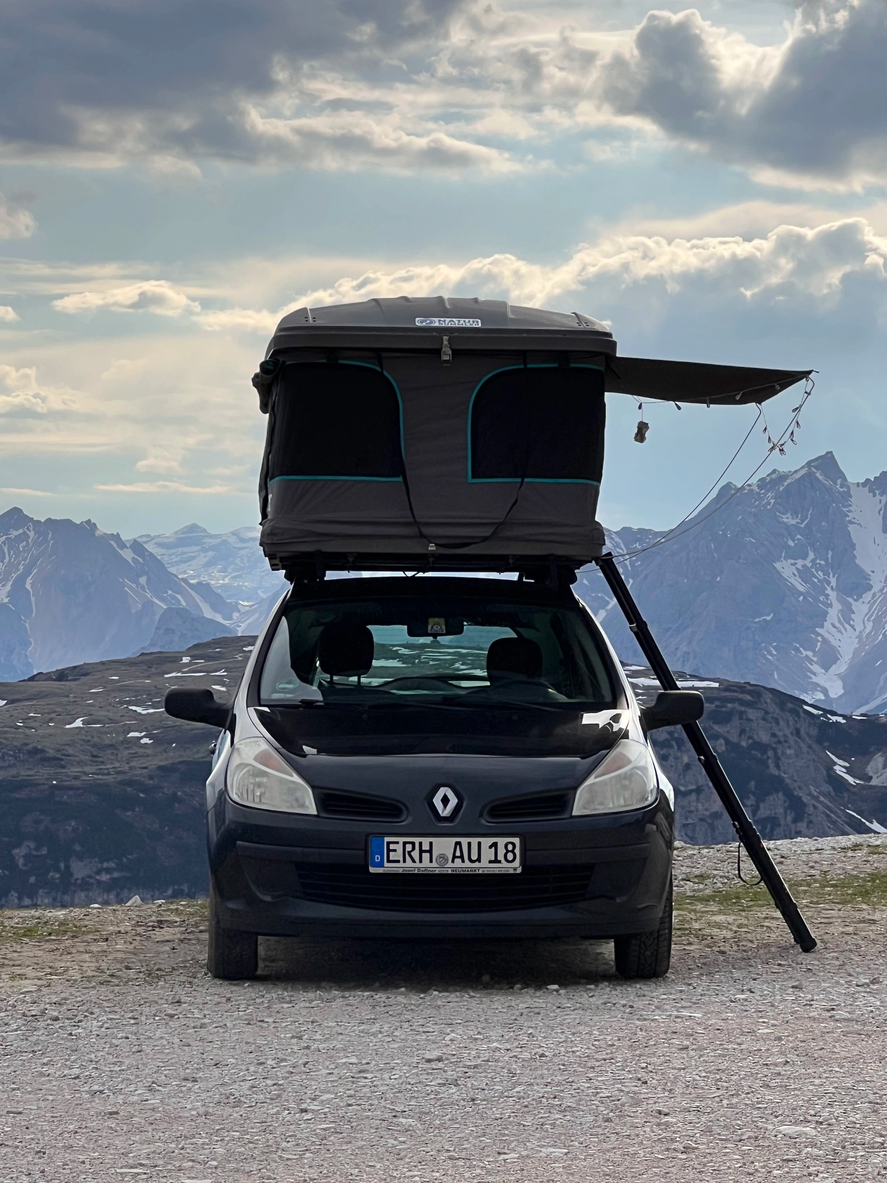 Frontalaufnahme von einem blauen Renault mit einem Dachzelt. Im Hintergrund sieht man eine Gebirgskette.